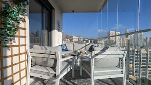 Un balcón con sofá y sillas, una mesita con una bandeja, y vista a la ciudad a través de amplias ventanas.