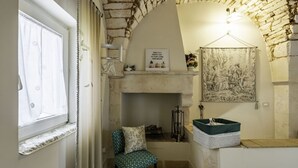 A cozy room with a stone wall, a fireplace, a chair, a pet bed, and a window with curtains. - A Casa di Marilù by Rentbeat (Martina Franca)
