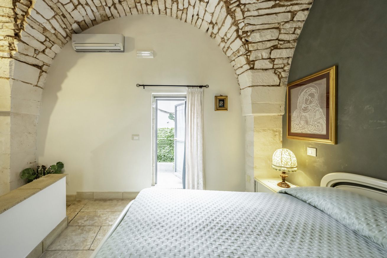 Ein Schlafzimmer mit steinbogiger Durchfahrt, einem Bett, einem Nachttisch mit Lampe und einem gerahmten Bild an der Wand.