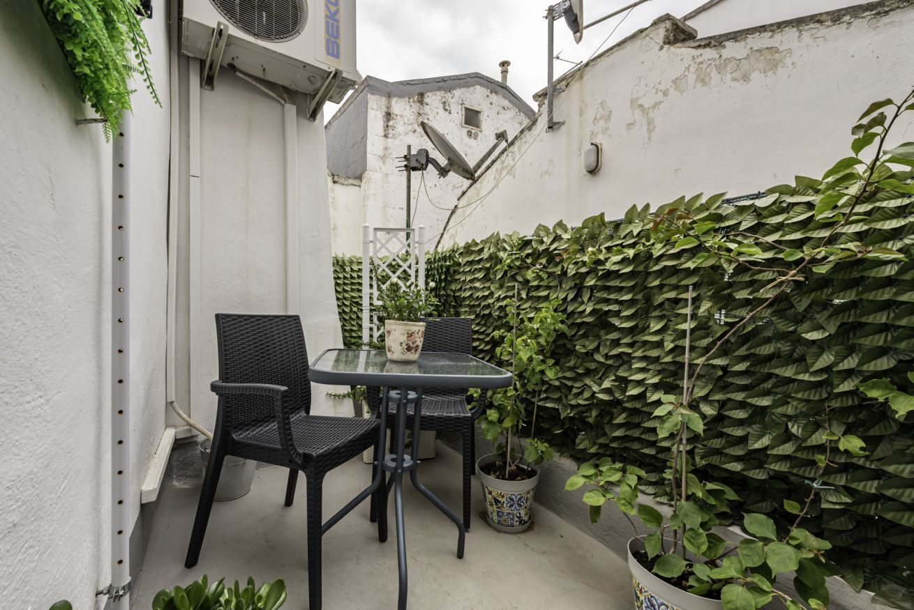 Un petit espace extérieur aménagé avec une table et des chaises, entouré d’un mur blanc et d’une haie verte.