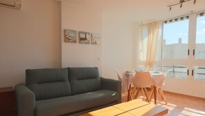 Ein Wohnzimmer mit grauem Sofa, einem Holztisch und einem Essbereich mit kleinem Tisch und Stühlen. An der Wand hängen drei Gemälde und ein Fenster mit Vorhängen.