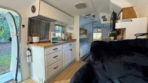 A compact RV interior with a kitchenette, a bed, and a dark-colored sofa.