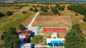 Vue aérienne d’un complexe sportif comprenant un court de tennis, des panneaux solaires, une piscine et un bâtiment avec un toit rouge.