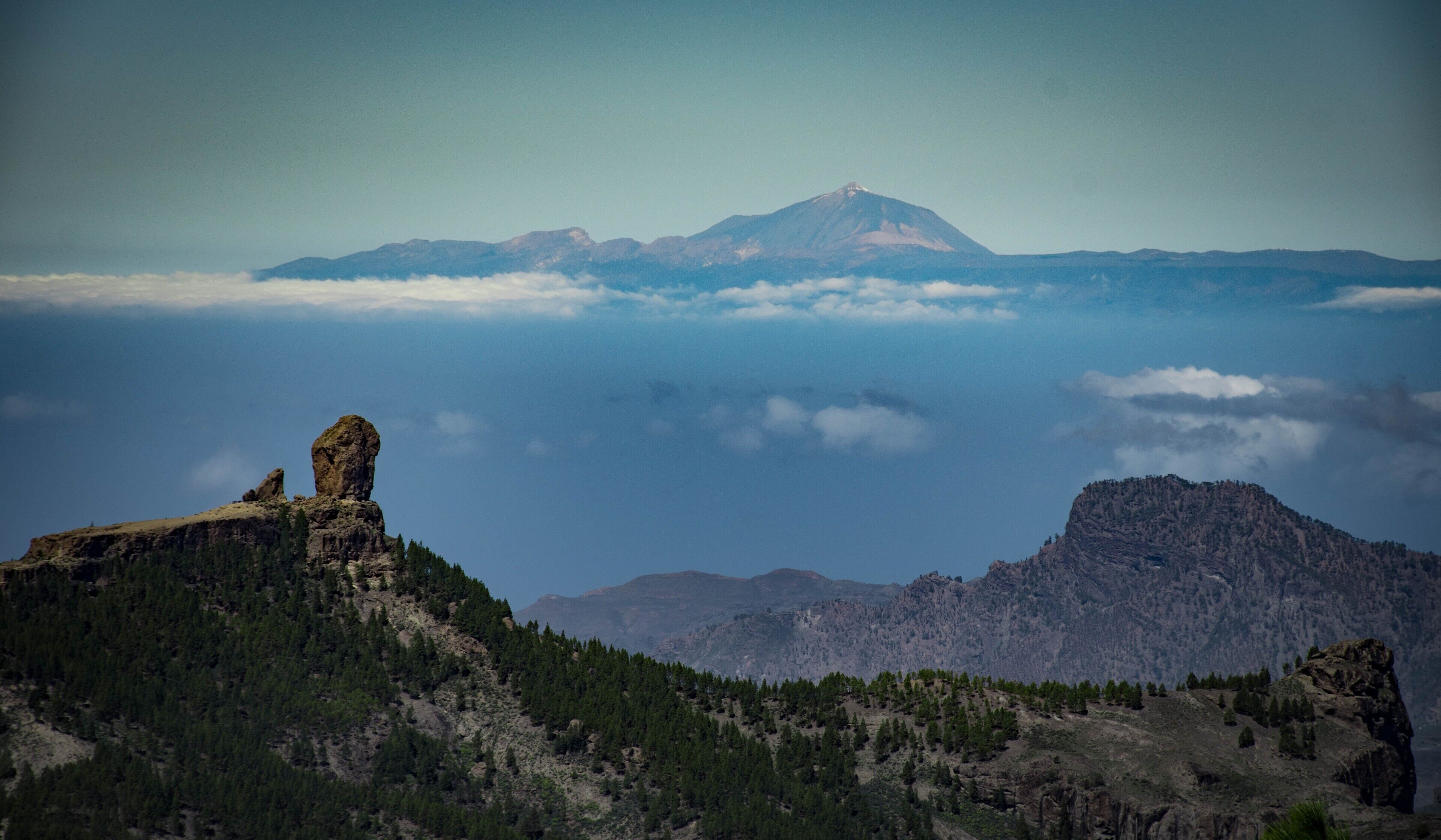 Un paysage montagneux avec une formation rocheuse imposante au premier plan, une zone boisée et un sommet montagneux lointain.