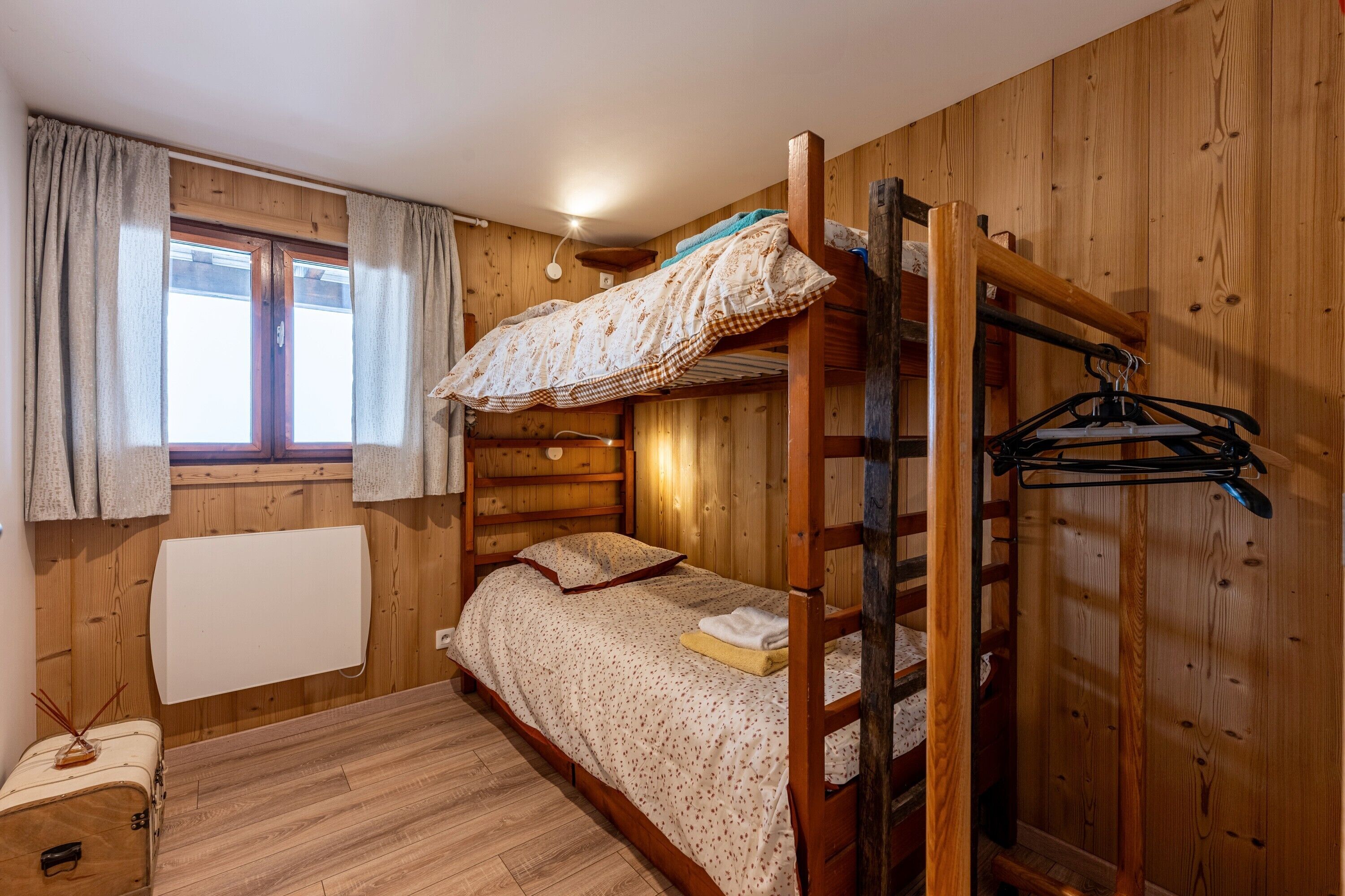 A bunk bed room with wooden walls, a window with curtains, a radiator, and a small table with a suitcase.