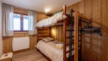 A bunk bed room with wooden walls, a window with curtains, a radiator, and a small table with a suitcase.