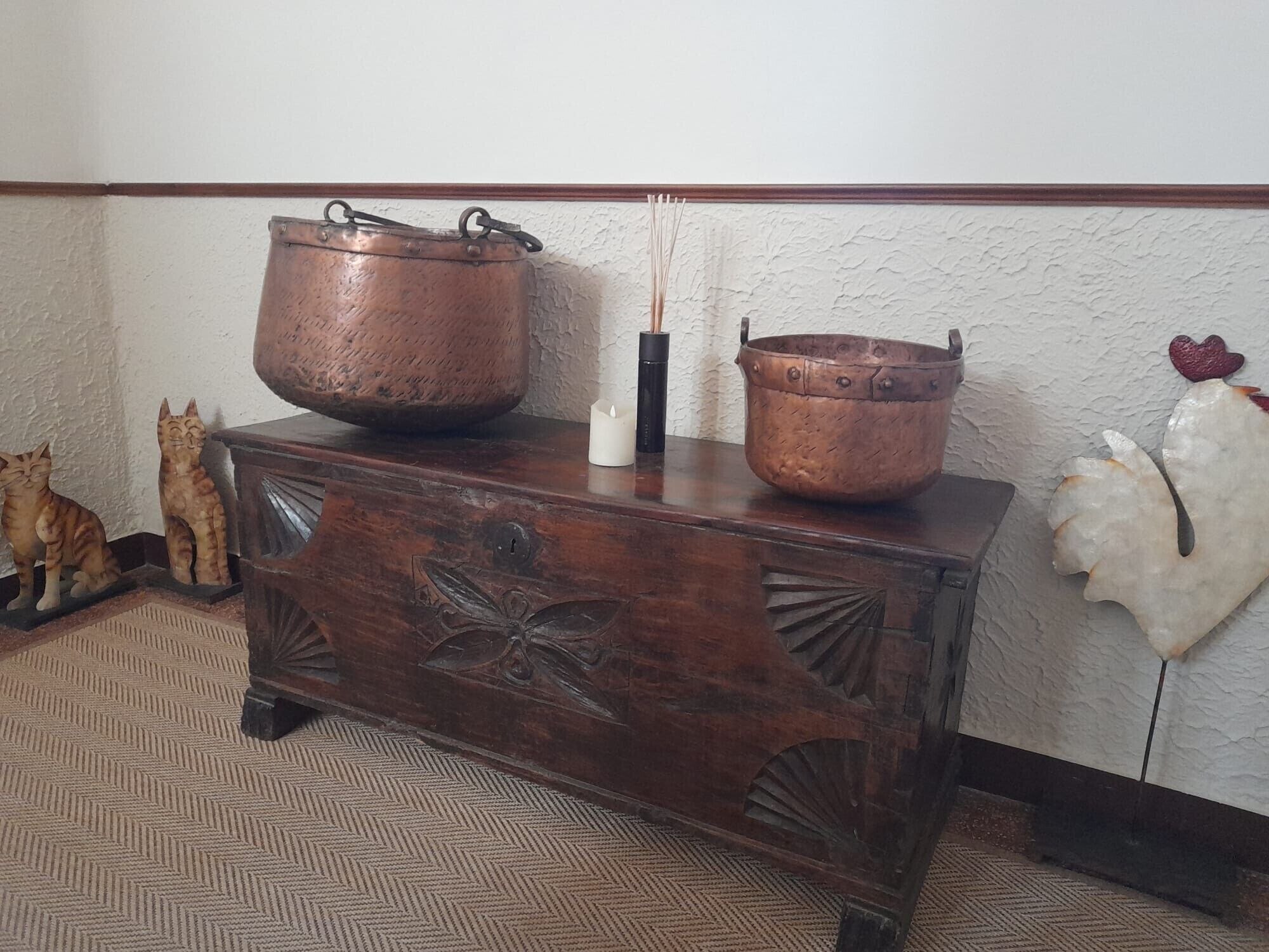 A wooden chest with two copper pots on top, a white candle, and a carved wooden cat figurine.