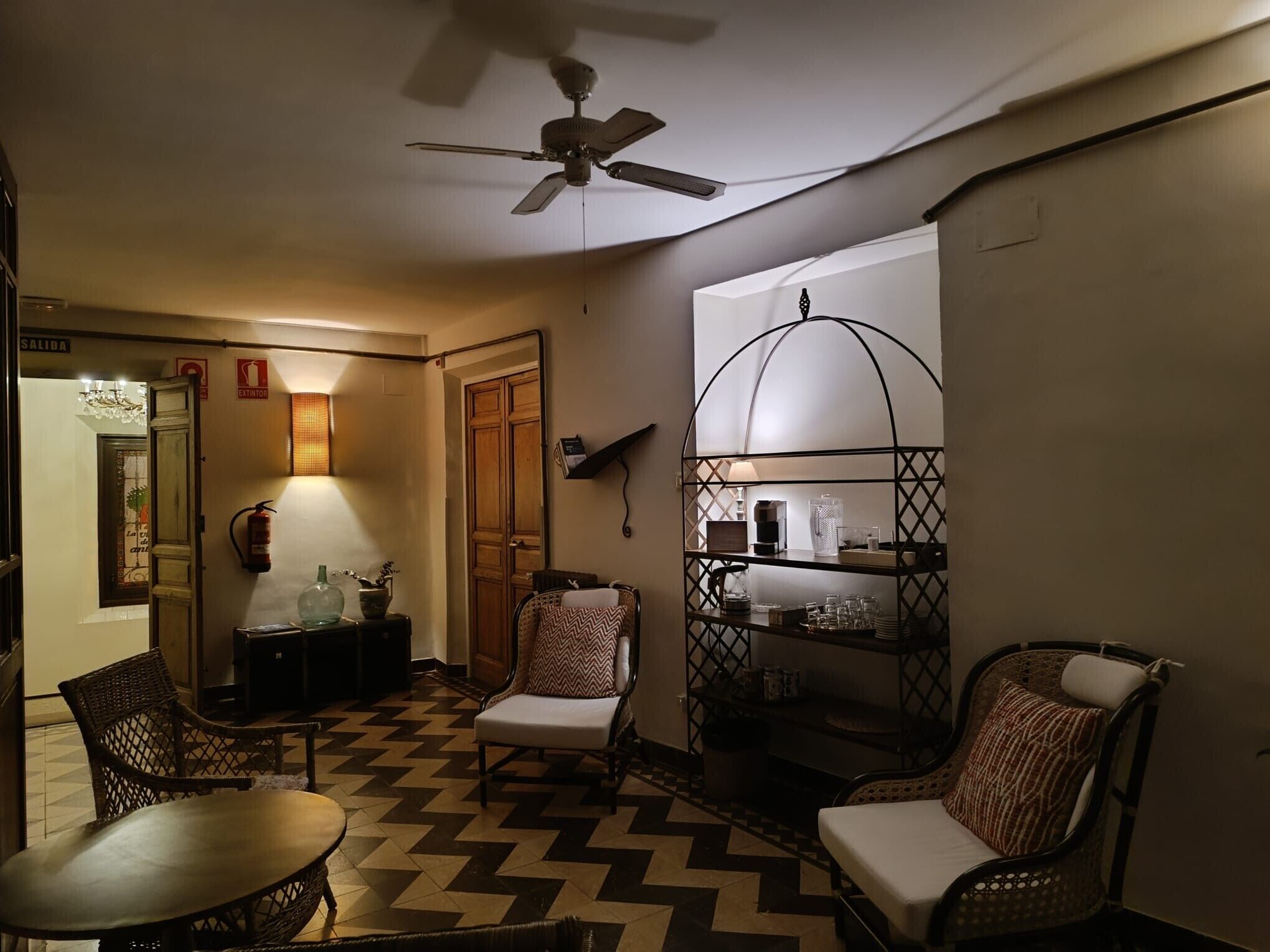 A room with a ceiling fan, a chair, a table, a shelf with items, and a patterned floor.