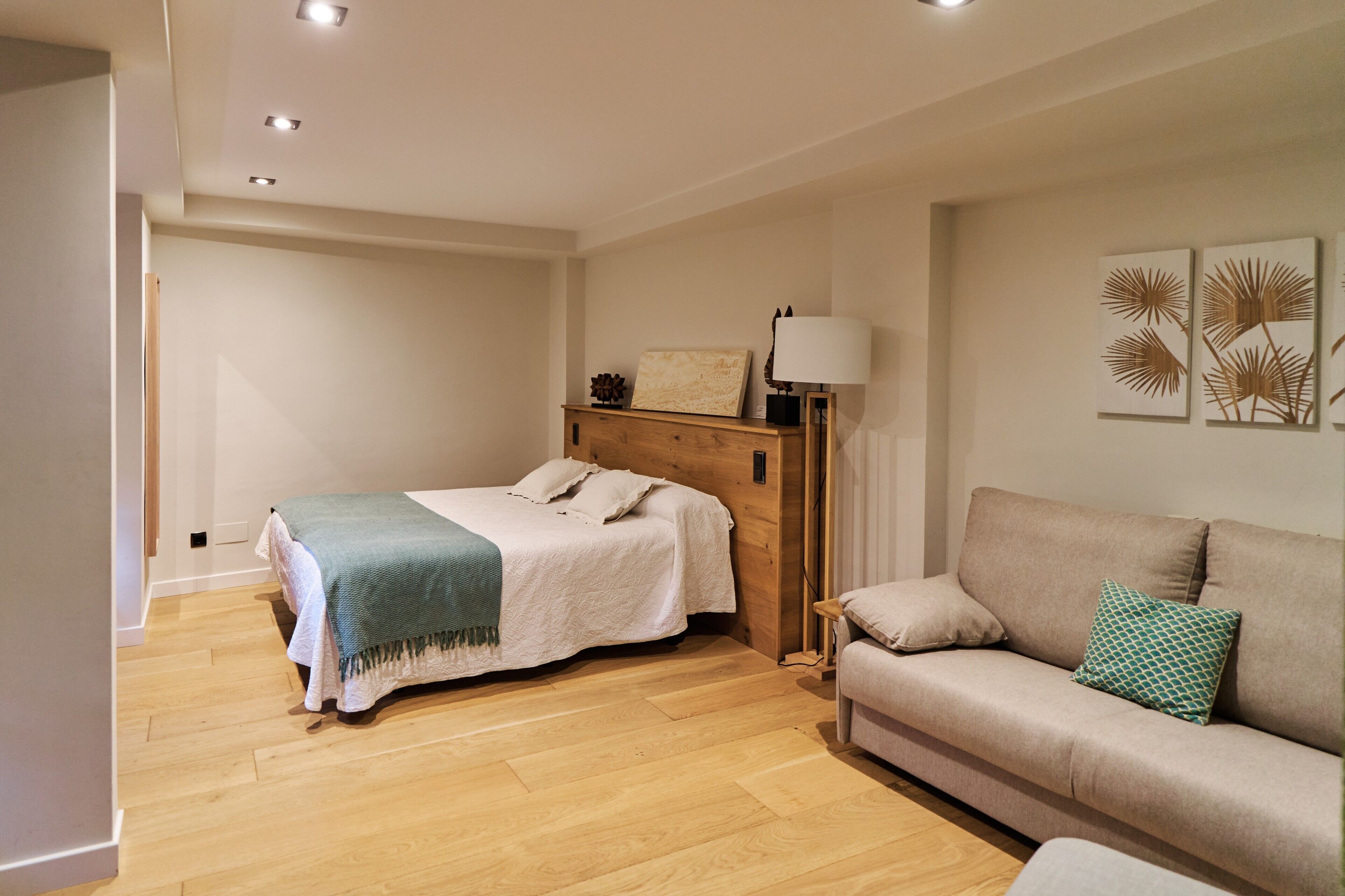 A bedroom with a bed, a wooden headboard, a bedside table with a lamp, a sofa, and wall art.
