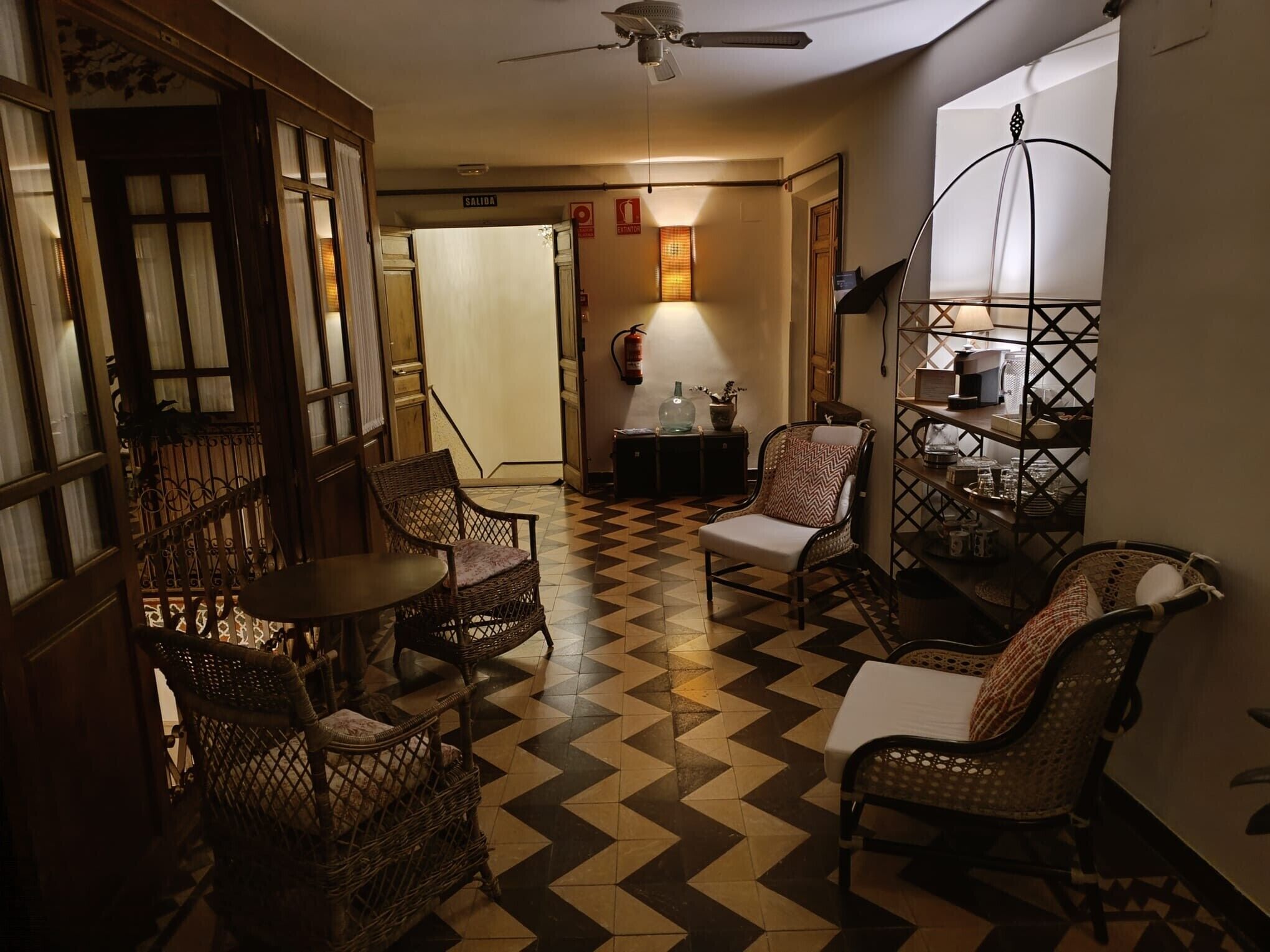 Ein Zimmer mit Schachbrettmuster-Boden, Korbmöbeln, einem Deckenventilator und einem an der Wand befestigten Regal mit Gegenständen.