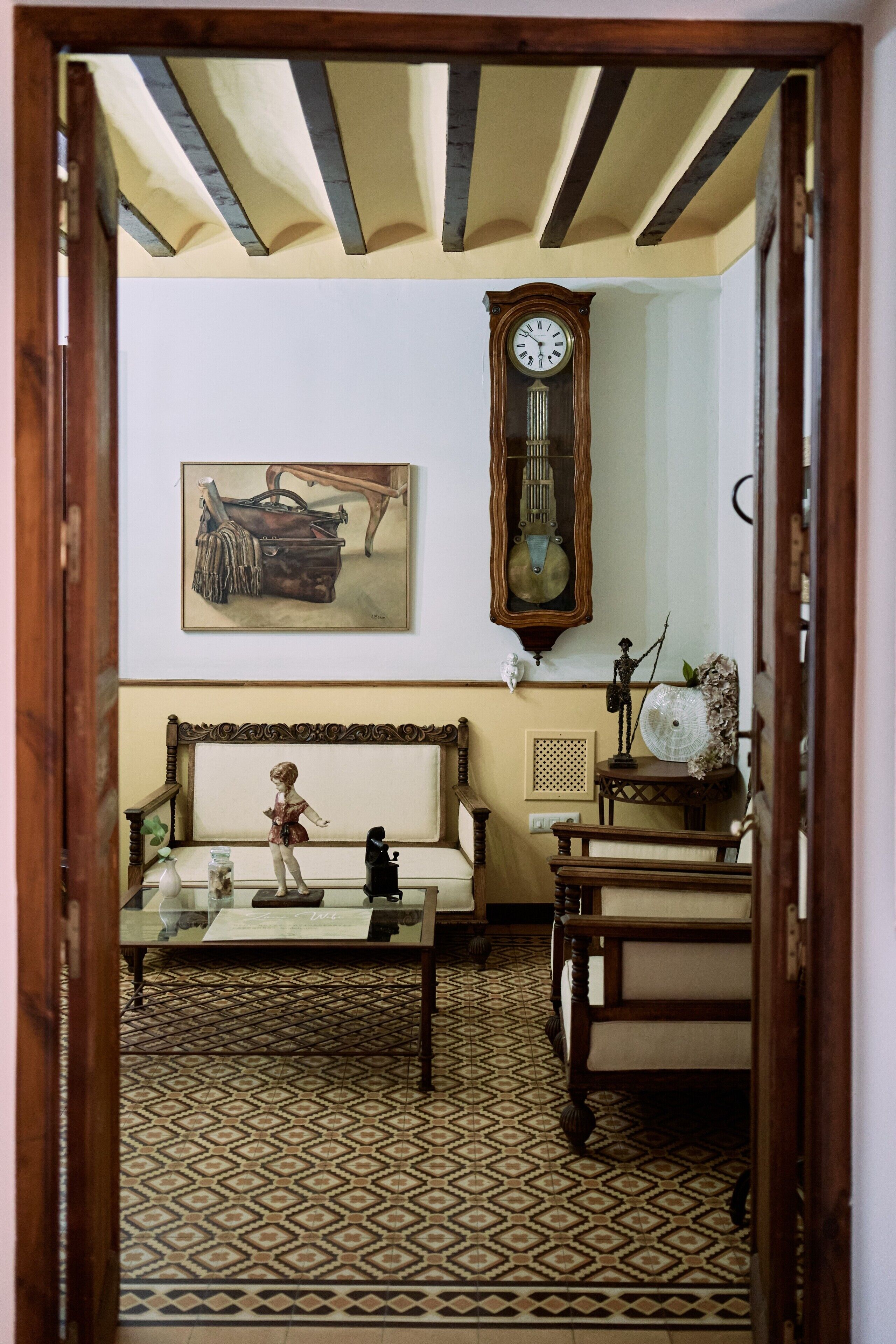 A room with a patterned floor, a wooden bench, a table with a figurine, a clock, and a painting on the wall.