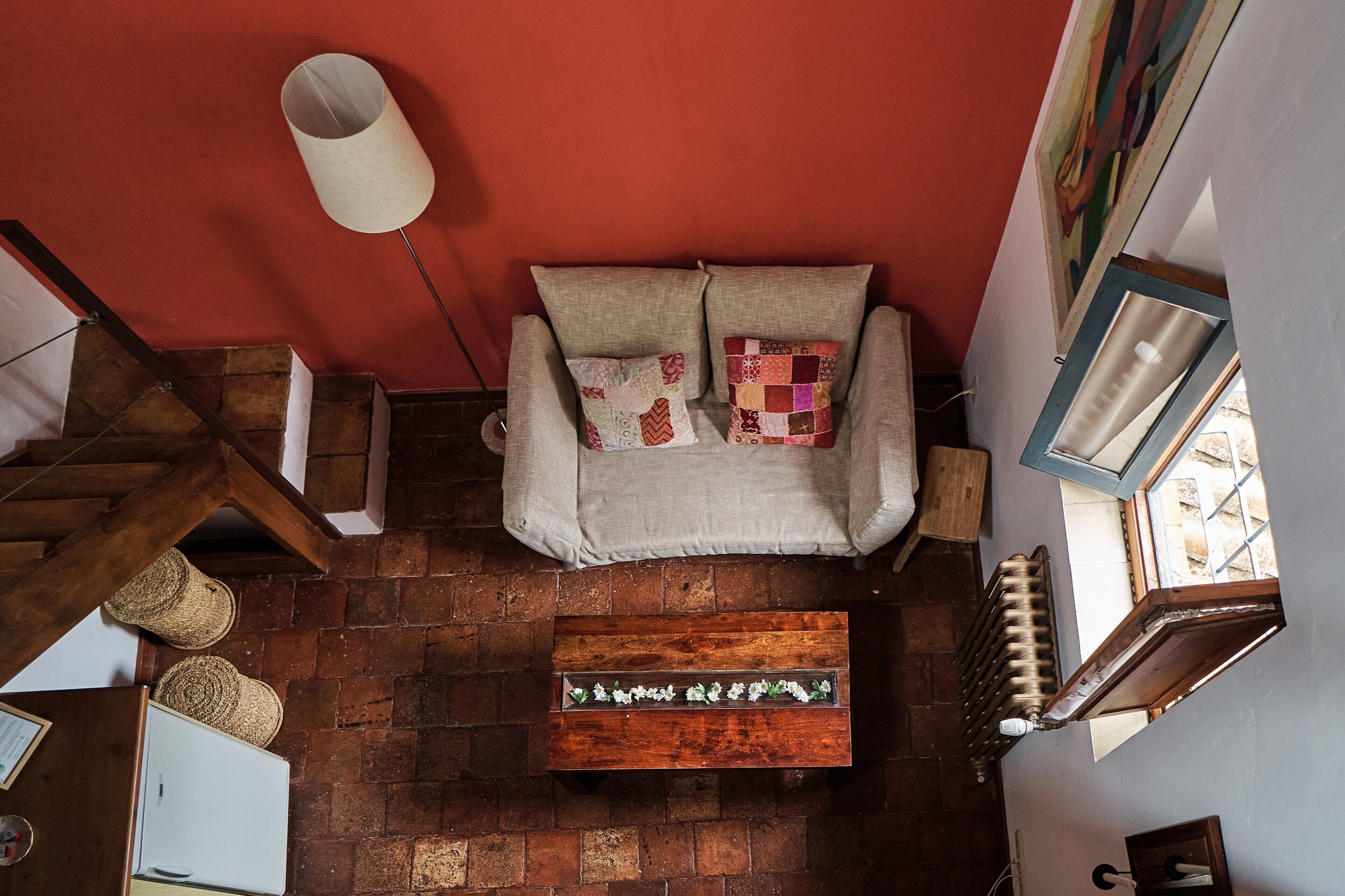 A room with a red wall, a sofa with patterned cushions, a wooden coffee table with flowers, a wooden staircase, and a radiator.