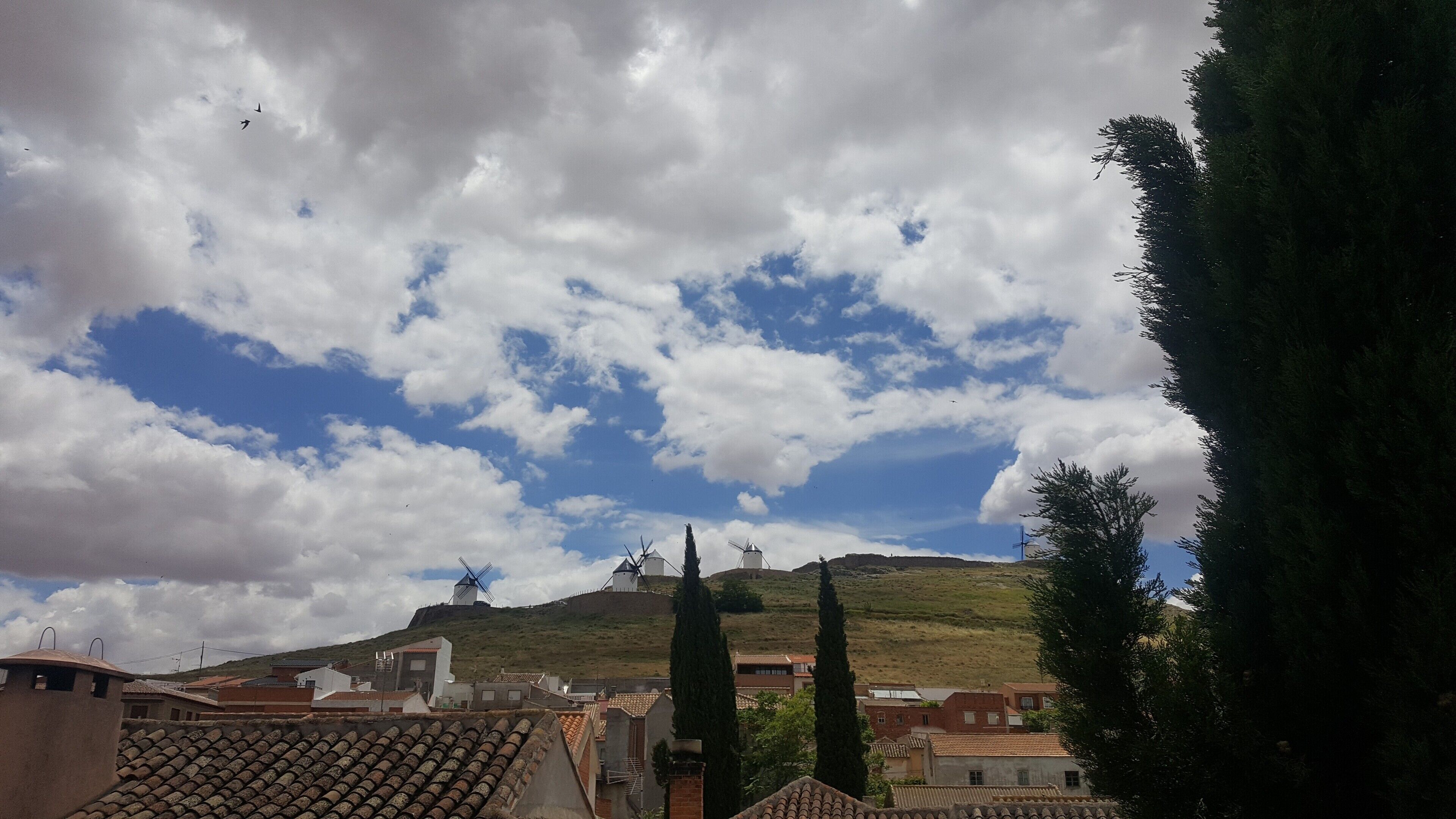 Une ville avec des moulins à vent perchés sur une colline, des bâtiments aux toits de tuiles et un ciel dégagé parsemé de nuages.