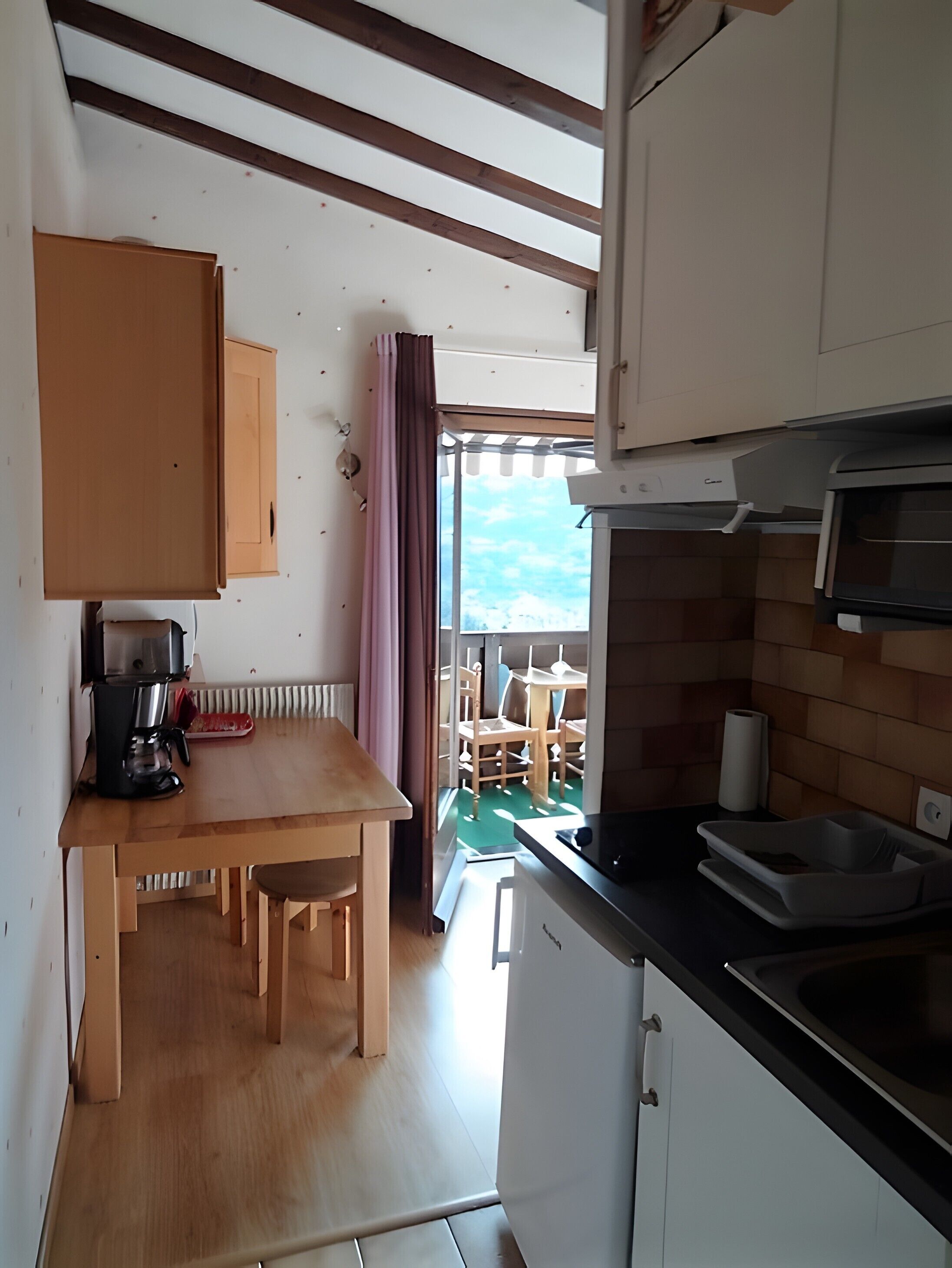 Una cucina compatta con mobili in legno, una macchina per il caffè e vista su un'area salotto esterna tramite una porta.