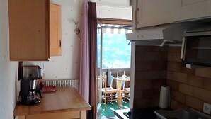 A compact kitchen with wooden cabinets, a coffee maker, and a view through a door to an outdoor seating area. - Studio Apartment 'Saint-Gervais' with Mountain View, Shared Pool and Private Garden (Saint-Gervais-les-Bains)