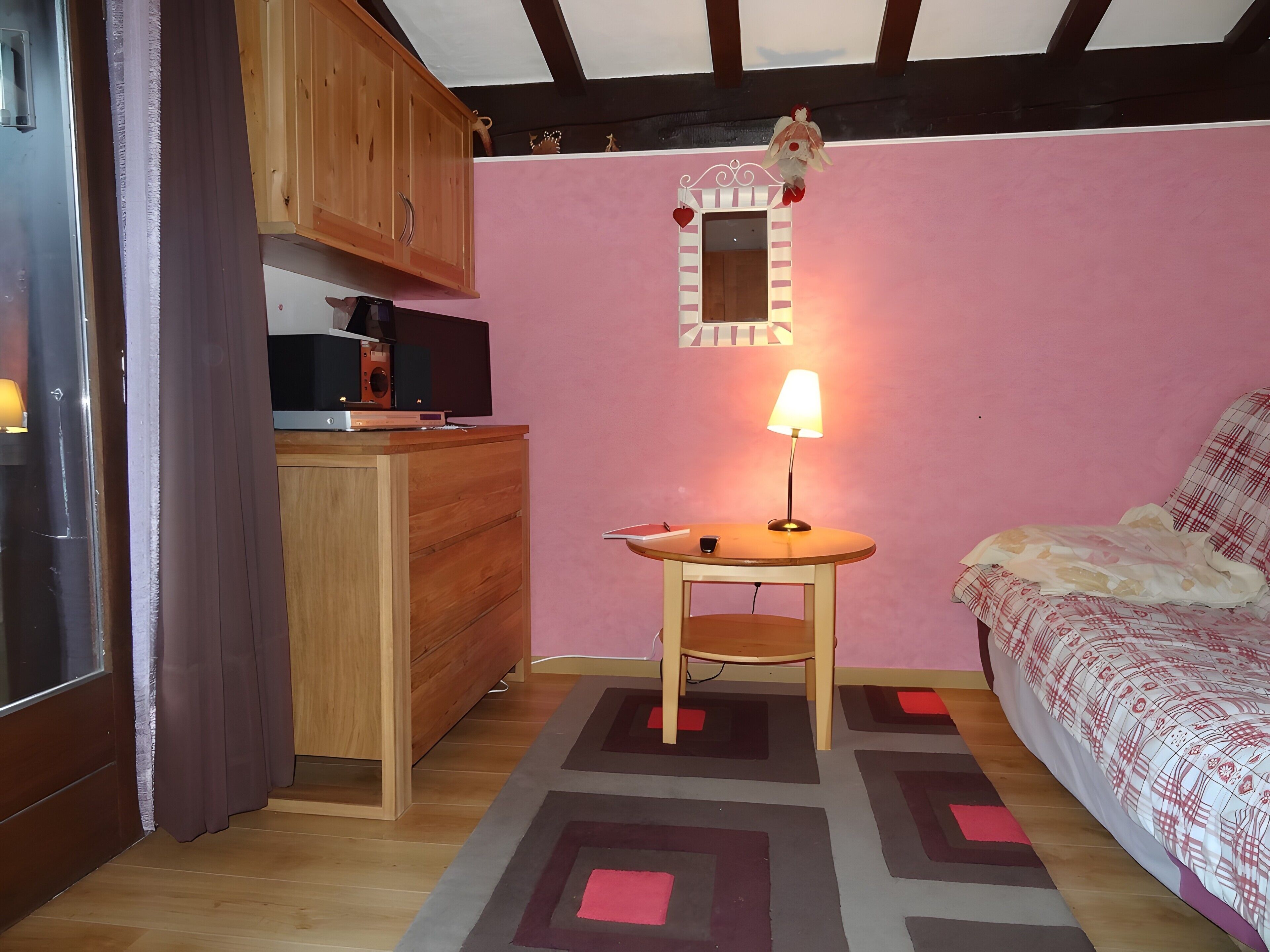 Una stanza piccola con un letto, una scrivania con una lampada, uno specchio e un angolo cottura con pensili e un televisore.