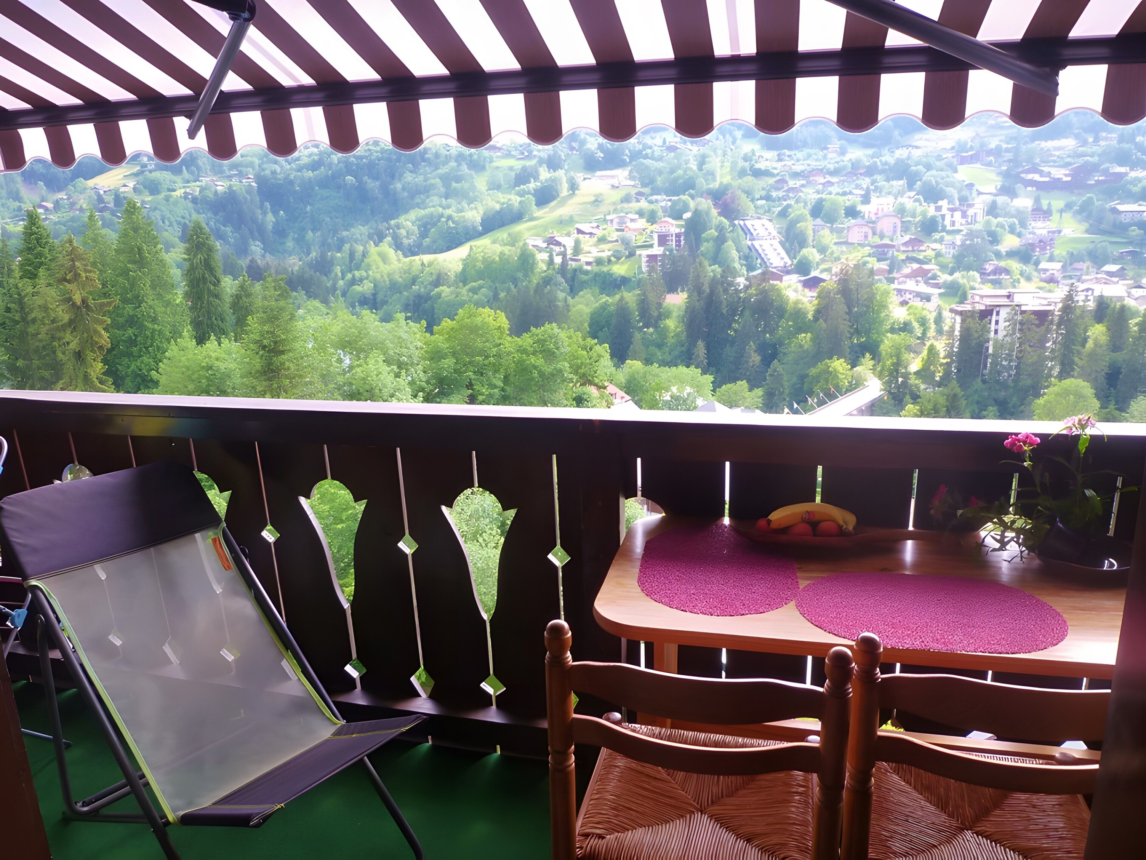 Un balcone con vista su una valle verde, un tavolo con un sottotavolo viola e due sedie in vimini.