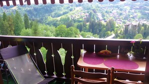 A balcony with a view of a green valley, a table with a purple mat, and two wicker chairs. - Studio Apartment 'Saint-Gervais' with Mountain View, Shared Pool and Private Garden (Saint-Gervais-les-Bains)