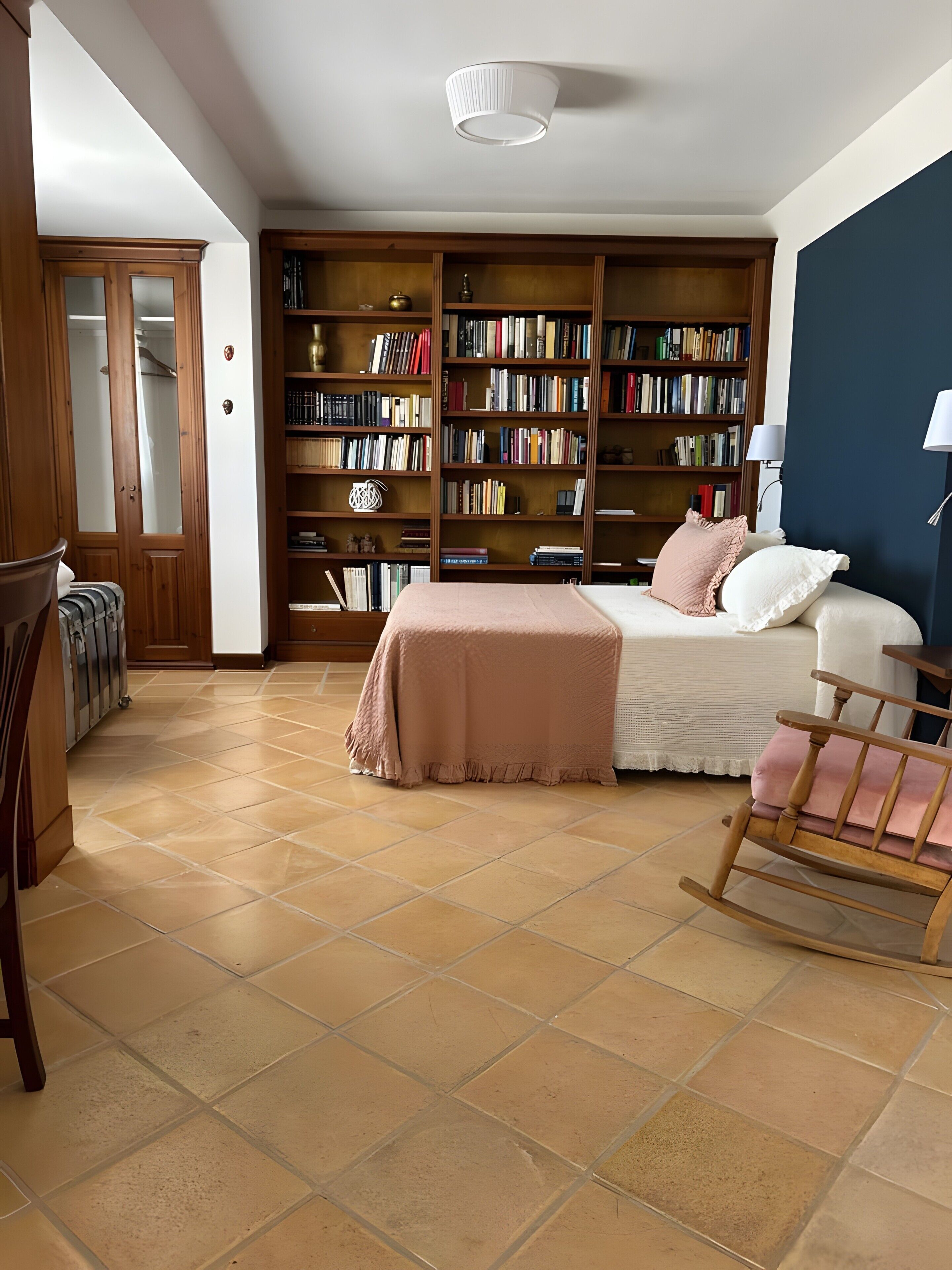 Una habitación con una cama, un sillón mecedora, una estantería y suelo de baldosas.