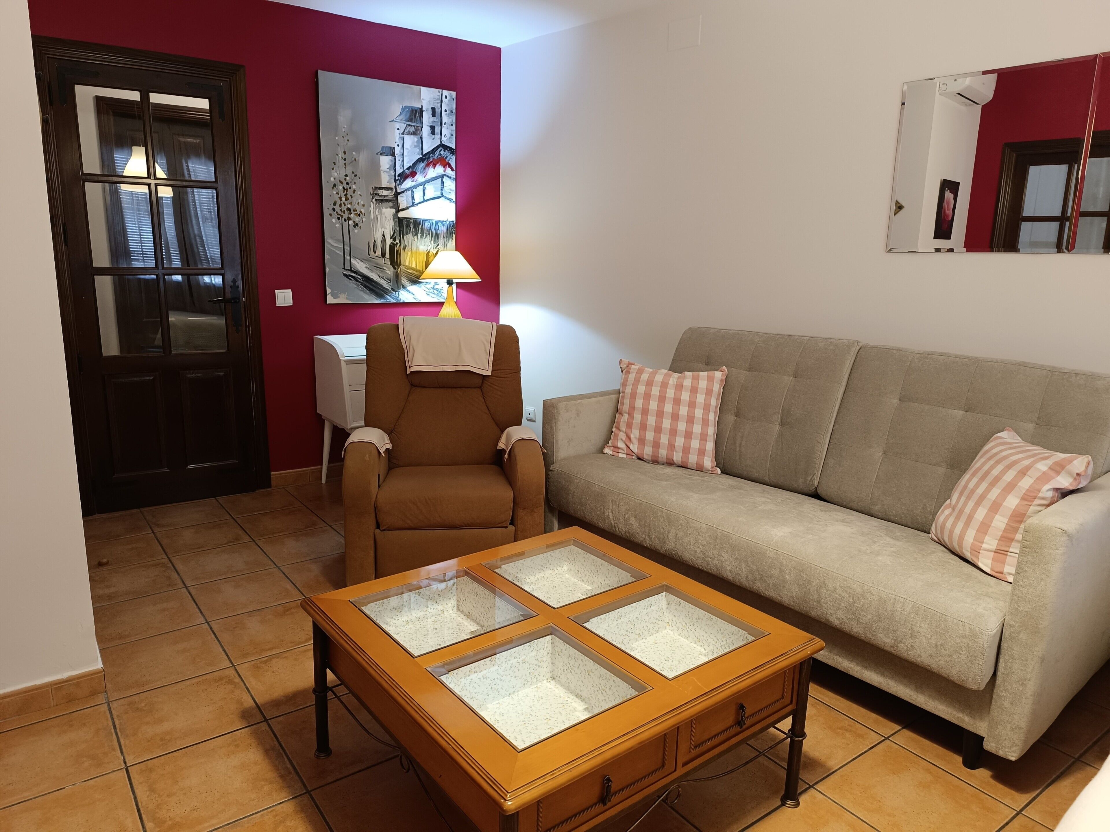Una sala de estar con un sillón marrón, un sofá beige con almohadones cuadriculados, una mesa de centro con superficie de vidrio, un cuadro en la pared y una puerta con ventanal.
