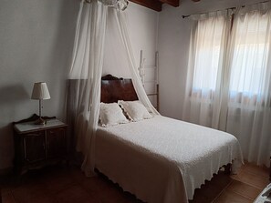 A bedroom with a canopy bed, a bedside table, and a window with sheer curtains.