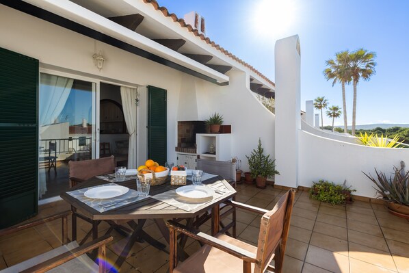 An outdoor patio with a table set for dining, a grill, and potted plants.