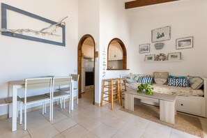 A living room with a sofa, coffee table, and chairs. There is a kitchen visible through an archway.