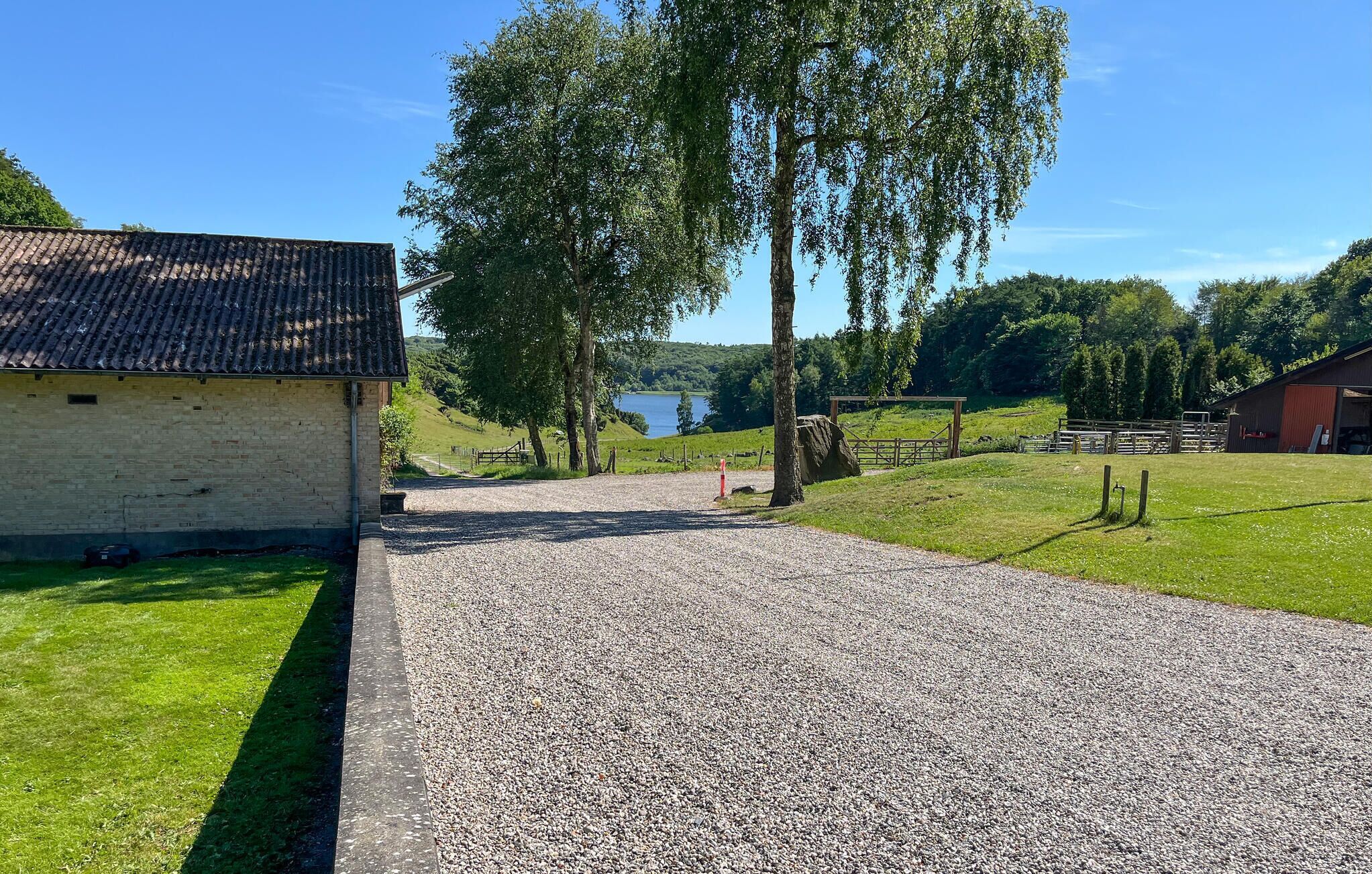 A gravel driveway leading to a building with a tiled roof, surrounded by greenery and a view of a lake.