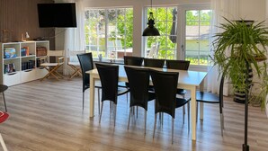 Uma área de refeições com uma mesa e cadeiras, uma estante de livros, uma televisão e uma grande janela com vista para o verde.