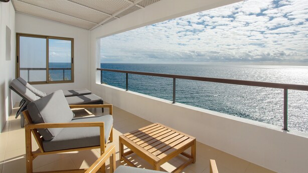 A balcony with two lounge chairs and a wooden table, overlooking a body of water with a clear sky.