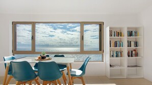 A room with a dining table, chairs, a bookshelf, and a large window with a view of the sea.