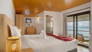 A hotel room with a bed, a wooden headboard, a bedside lamp, a bathroom visible through an open door, and a balcony with a view of the sea.
