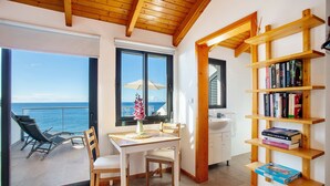 Een kamer met een balkon, een eettafel, stoelen, een boekenkast en uitzicht op zee.