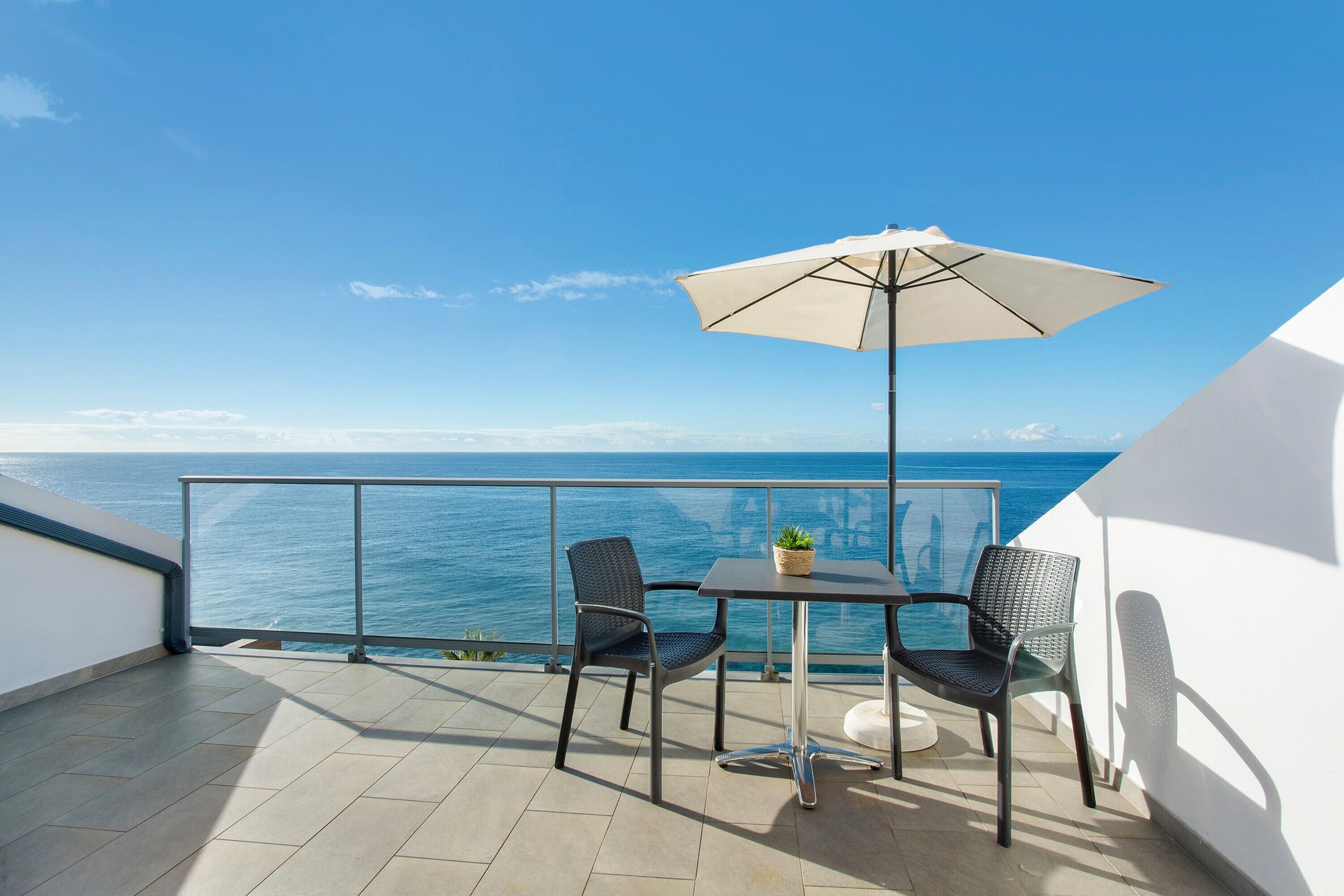 Un balcon avec une table, deux chaises, un pot de fleurs et un grand parasol.
