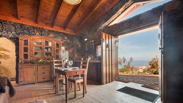 Un intérieur rustique avec un mur en pierre, des meubles en bois, une table à manger et une vue sur l’extérieur.