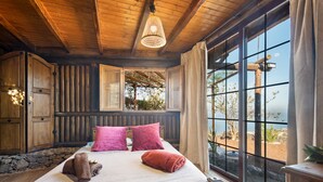 Une chambre à coucher de style cabane en bois, avec un grand lit, deux oreillers roses, une tête de lit en bois et une vue sur l’extérieur.