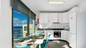 Cozinha moderna com armários brancos, micro-ondas e geladeira. Há uma área de refeições com mesa e cadeiras, e uma grande janela com vista para o mar.
