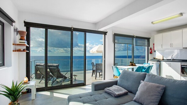 Uma sala de estar moderna com sofá, área de jantar e vista para o mar através de grandes janelas.