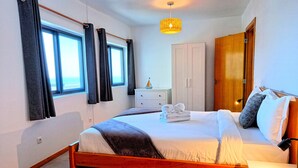 Quarto com cama de madeira, mesa de cabeceira, guarda-roupa, janela com cortinas e luminária suspensa.