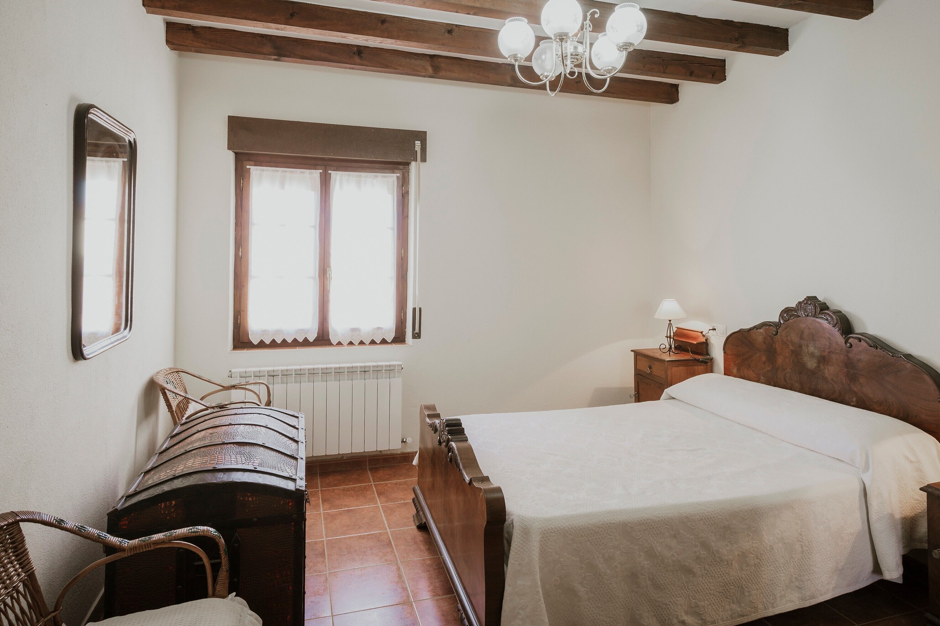 A bedroom with a wooden bed, a chair, a nightstand, a lamp, and a window with curtains.
