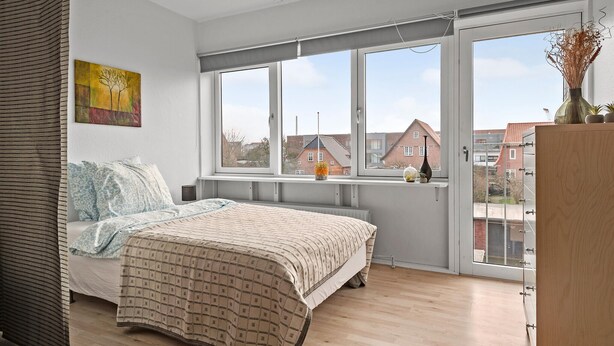 Une chambre avec une grande fenêtre, un lit, un radiateur et une vue sur les bâtiments environnants.