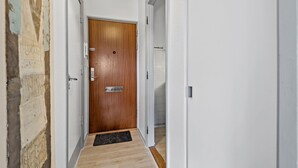 Un couloir avec une porte en bois, un paillasson noir et un mur blanc orné d’un motif texturé.