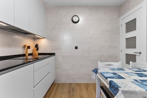 A compact kitchen with white cabinets, a black countertop, a wooden cutting board, a clock, and a table with a blue tablecloth. - Apartment 'Comarca Del Sella' with Mountain View and Wi-Fi (Arriondas)