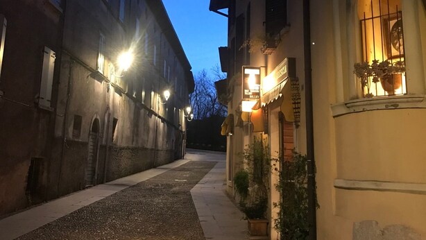 Una calle estrecha flanqueada por edificios, con un camino adoquinado y letreros iluminados.