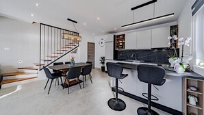 Una cocina moderna con encimera de mármol negro, horno empotrado y estante de madera. Cuenta con un comedor con una mesa redonda y cuatro sillas. Una escalera de madera con barandillas negras conduce a otro nivel.