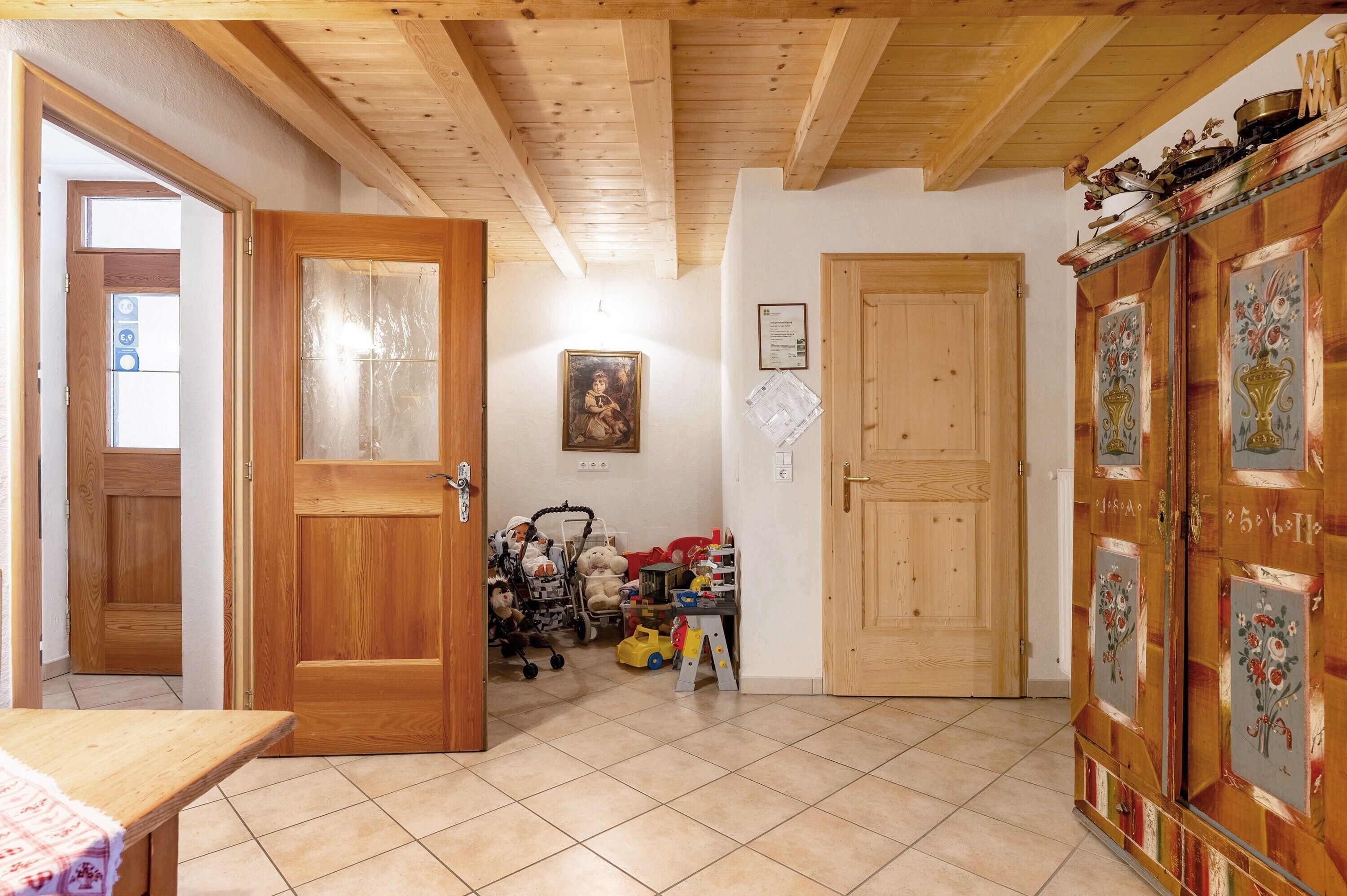 Un intérieur en bois avec un sol carrelé, une porte en bois, un tableau accroché au mur et un meuble orné de motifs floraux.