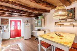 A rustic kitchen with exposed wooden beams, a red door, a dining table with a patterned tablecloth, and a hanging wicker light fixture. - Holiday Home 'Tis Manas' with Sea View, Private Terrace and Wi-Fi (South Rhodes)
