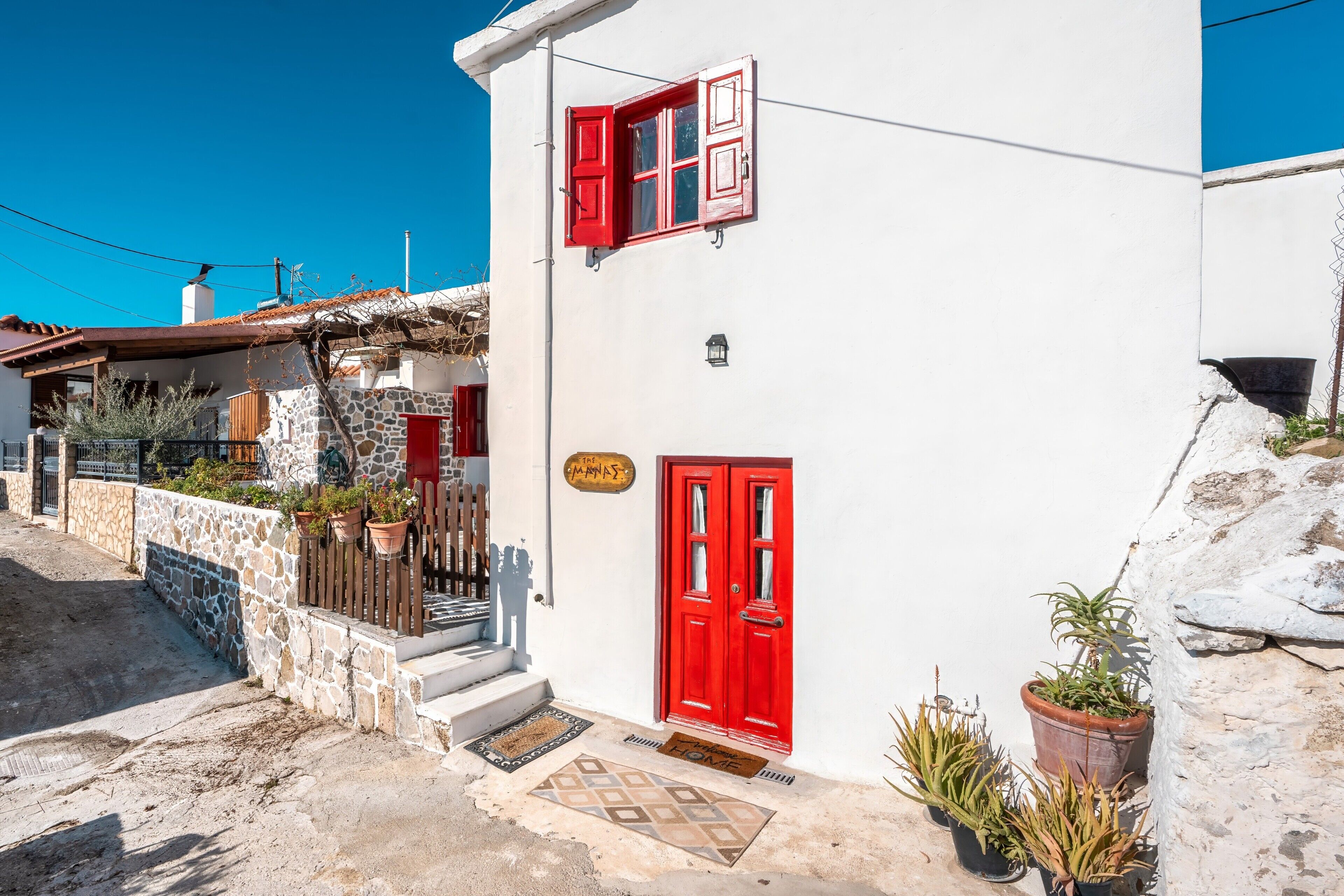 A white building with a red door and window shutters, stone walls, and potted plants.