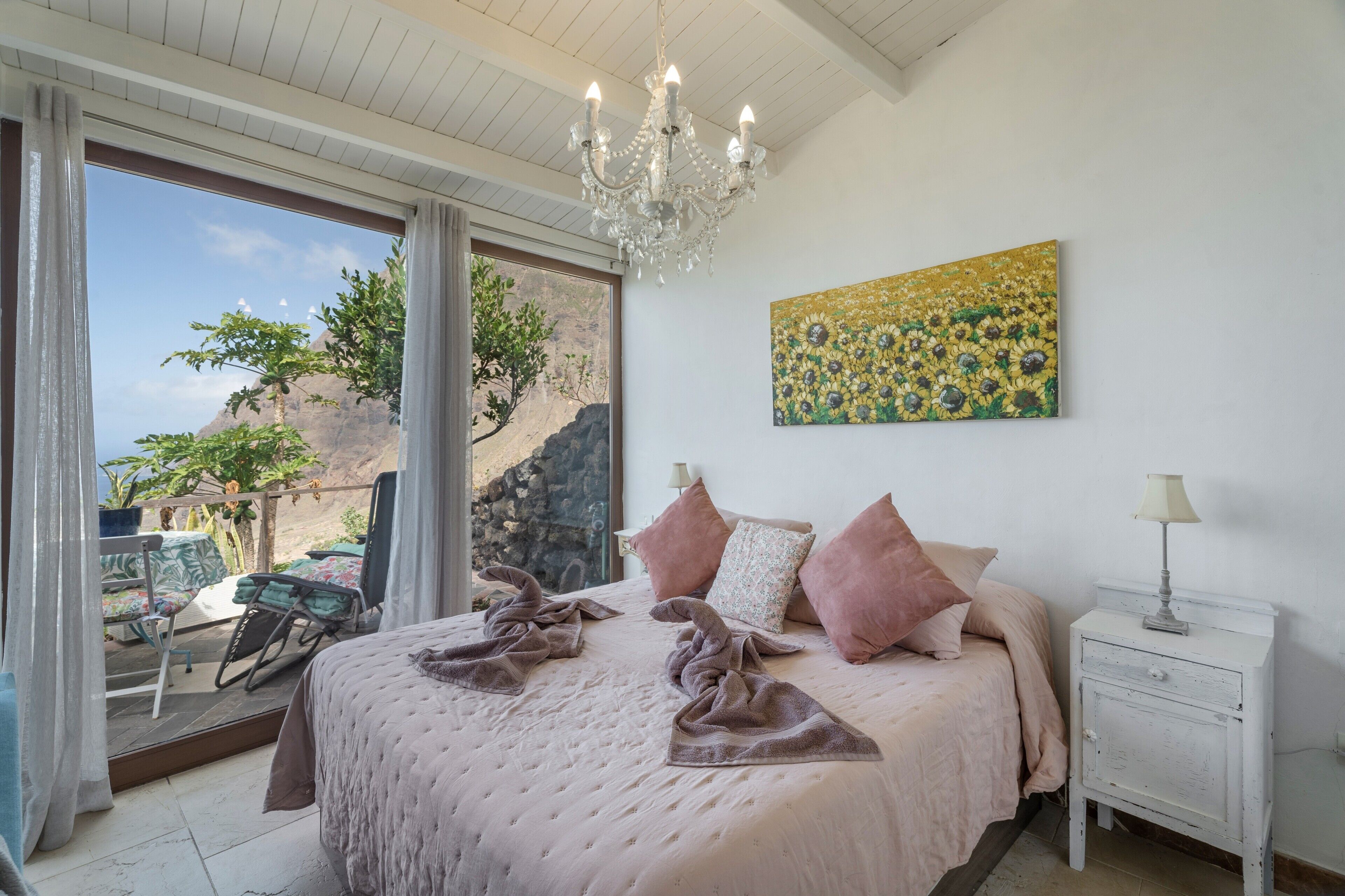 Ein Schlafzimmer mit einem großen Bett, einer Kristallleuchter, einem Gemälde und Blick auf eine felsige Landschaft durch ein Fenster.