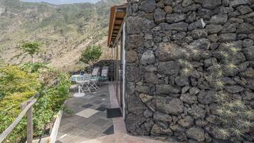Un muro de piedra con un patio, mobiliario exterior y montañas al fondo.